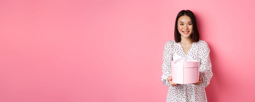 Beautiful asian woman wishing happy holidays, giving you gift in cute box, standing against pink background.