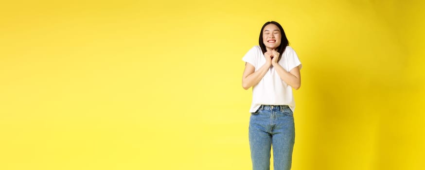 Full size portrait of excited asian girl winning and celebrating, achieve success, smiling cheerful, standing over yellow background.