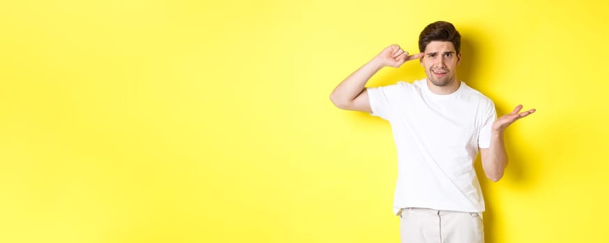 Confused and angry man pointing at head, scolding person for acting stupid, show crazy sign, standing over yellow background.