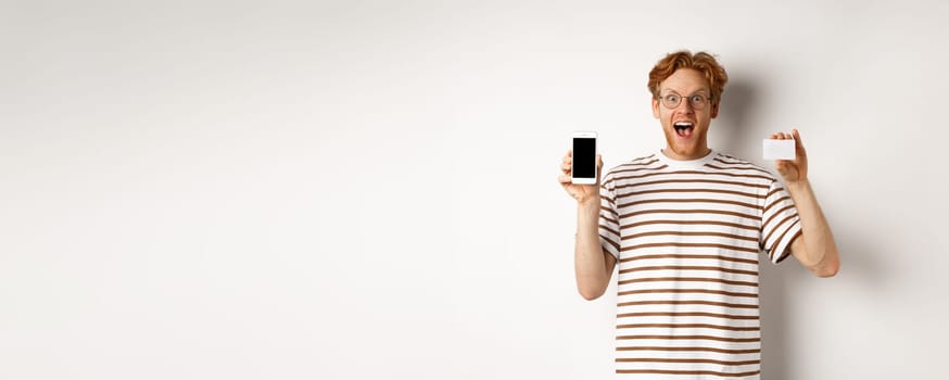 Shopping and finance concept. Young man showing blank mobile screen and plastic credit card, smiling at camera, white background.