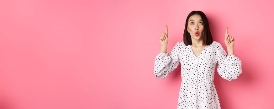 Cute korean girl in beautiful dress saying wow, looking and pointing fingers up, intrigued in promo offer, standing over pink background.