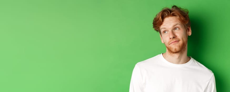 Smiling redhead man with messy haircut and beard tilt head, looking left with pleased face, standing over green background.