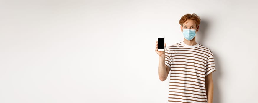 Covid-19, online doctor concept. Smiling redhead man in face mask showing smartphone screen, standing over white background.