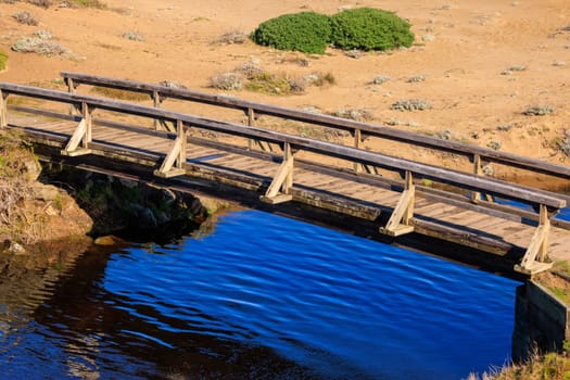 Wooden footbridge over blue water in dry desert sand dunes. High quality photo