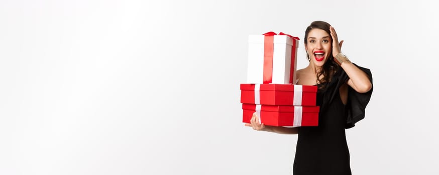 Celebration and christmas holidays concept. Excited and happy woman receive gifts, holding xmas presents and rejoicing, standing in black dress over white background.