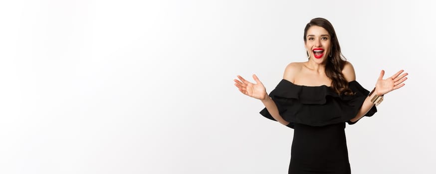 Beauty and fashion concept. Image of surprised and happy young woman in party dress reacting to good news, raising hands up and smiling amazed, white background.
