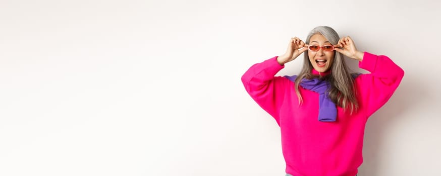 Fashionable asian senior woman trying new sunglasses, looking amazed and cheerful at camera, standing in trendy sweater against white background.