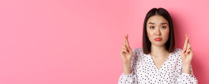 Beauty and lifestyle concept. Close-up of sad and hopeful asian woman making wish, cross fingers good luck and pouting, looking upset, standing over pink background.