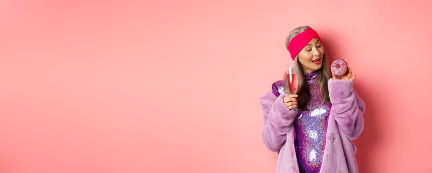 Fancy senior woman having fun, eating donut and drinking pink champagne, standing in purple faux fur coat and glittering dress, studio background.