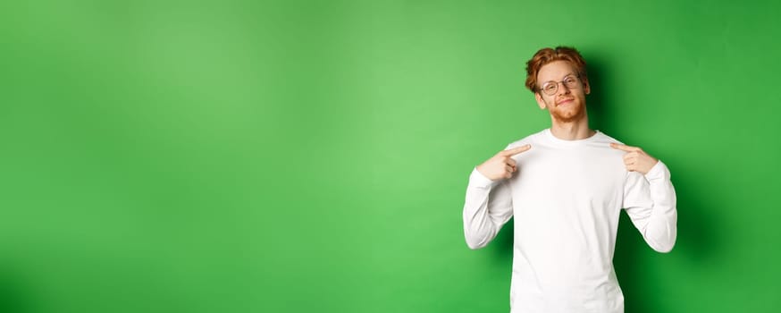 Confident and smug redhead man in glasses smiling, pointing at himself self-assured, standing over green background. Copy space