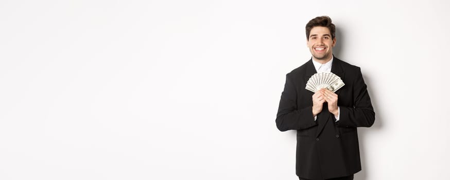 Portrait of happy and pleased handsome man in suit, hugging money and looking satisfied, standing over white background.