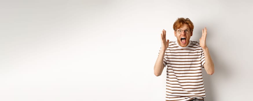 Angry young man with red hair shouting at camera, screaming and looking outraged, shaking hands, standing over white background.