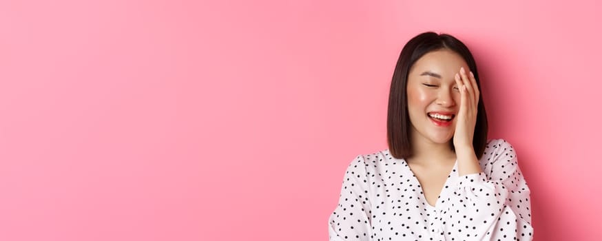 Beauty and lifestyle concept. Close-up of happy asian female laughing, looking happy and showing genuine emotions, standing over pink background.