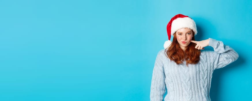 Winter holidays and Christmas Eve concept. Sassy teenage girl with red hair, wearing santa hat, enjoying New Year, poking her cheek and looking confident at camera.