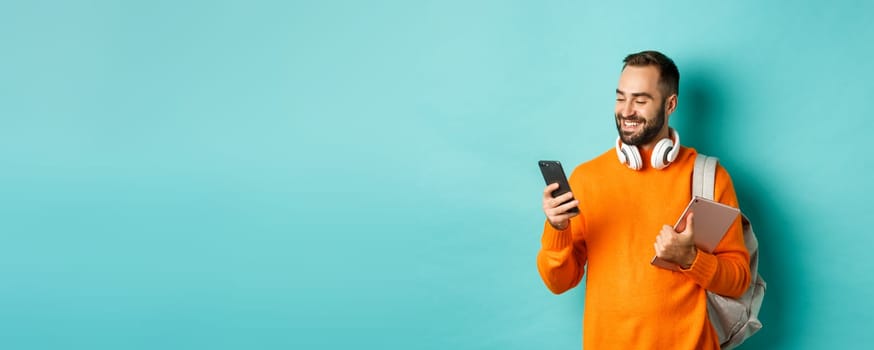 Handsome man student with headphones and backpack, holding digital tablet, reading message on mobile phone, standing against light blue background.