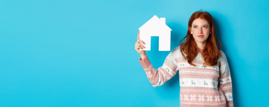 Real estate concept. Image of cute redhead girl looking curious at paper house model, thinking of buying property, looking up at copy space, blue background.