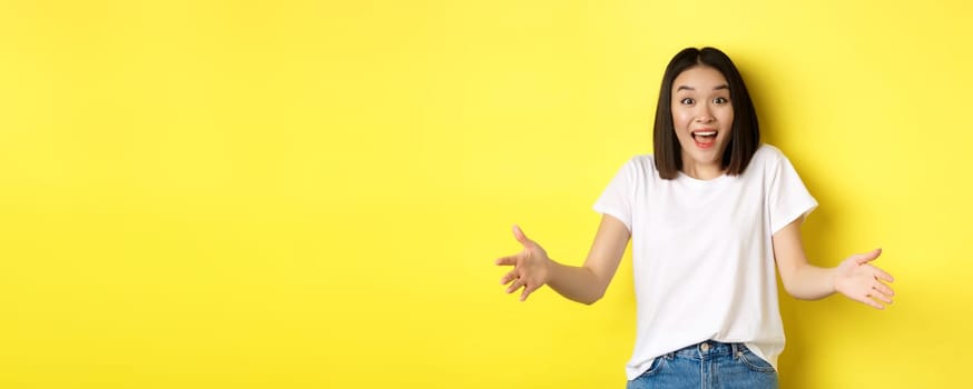 Excited young asian woman spread out hands as if holding large, big object, showing size and look amazed, standing over yellow background.