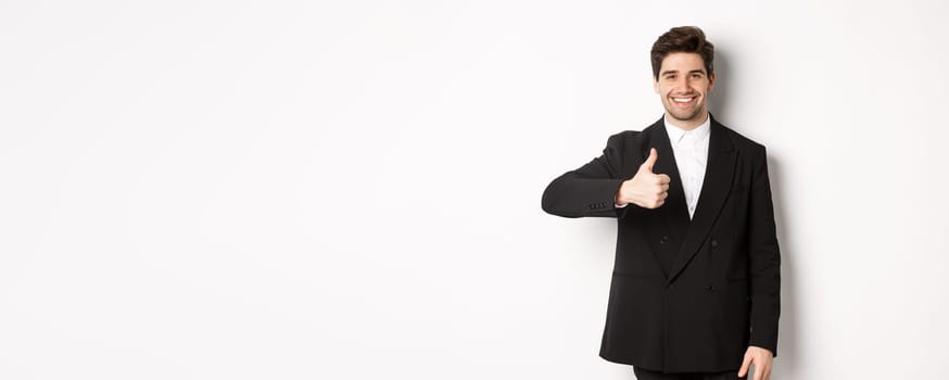 Portrait of handsome and confident male realtor, showing thumb-up and smiling, guarantee quality and recommending company, standing over white background.