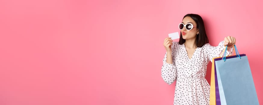 Beautiful asian woman in sunglasses going shopping, holding bags and kissing credit card, standing over pink background.