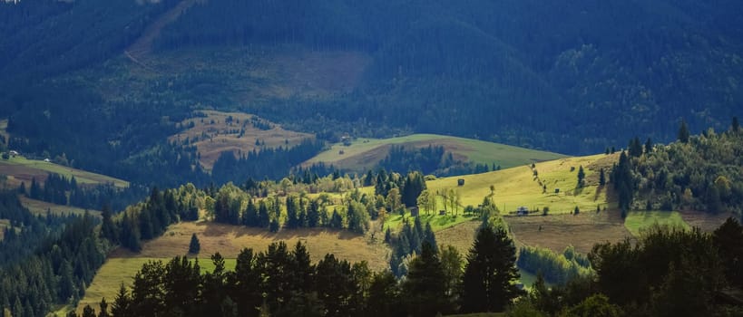 Wonderful mountain land with forest and small houses in sunlight. Scenic view to vast expanses of rolling hills and great mountain ridge on horizon. Immense distances of wavy relief in evening light