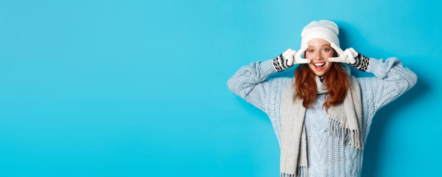 Winter and holidays concept. Cute redhead teen girl in beania, gloves and sweater showing peace sign, looking left at camera and wishing merry christmas, standing against blue background.