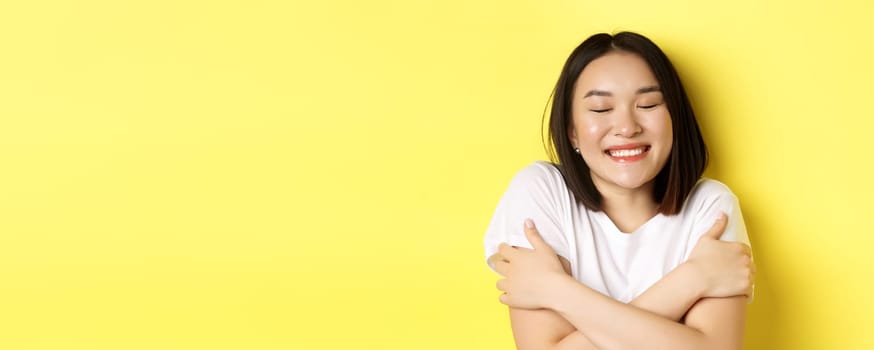Close up of romantic asian girl hugging herself and dreaming, close eyes and smile while imaging something tender, standing over yellow background.