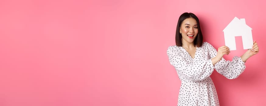 Real estate. Beautiful asian woman demonstrating paper house model, looking at camera confident, advertising home for sale, standing over pink background.