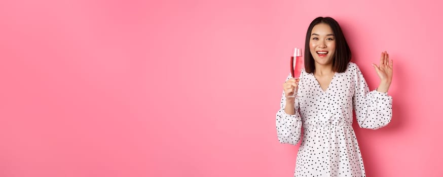 Celebration. Beautiful asian woman drinking champagne and smiling, standing in dress over pink background.