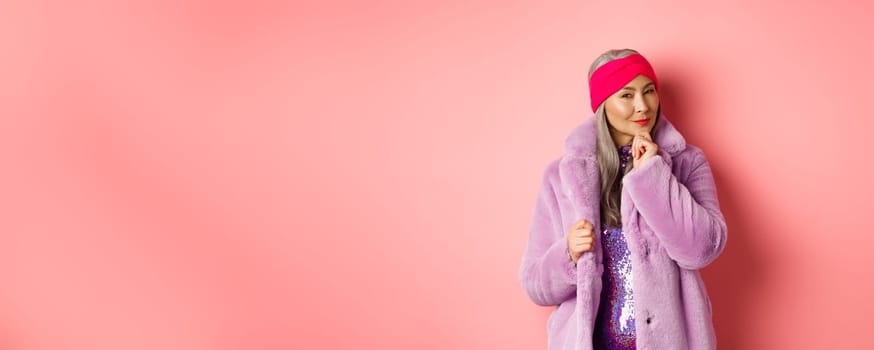 Fashion and shopping concept. Stylish old asian lady in purple fake-fur coat looking intrigued, interested with promotion, smiling and thinking, pink background.