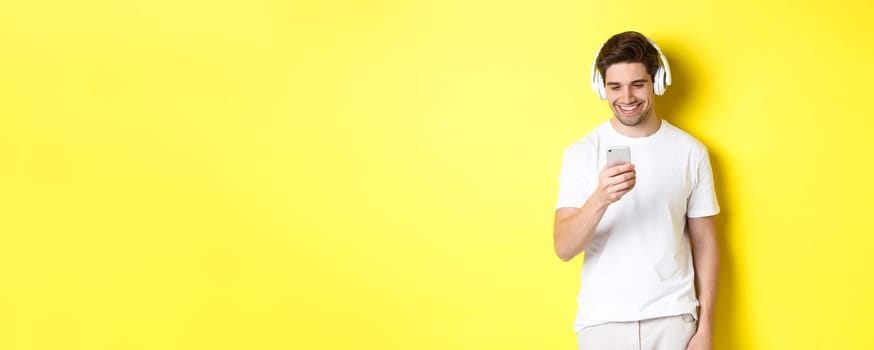 Young man in headphones reading message on smartphone, smiling, listening music in earphones and picking song in app, standing over yellow background.