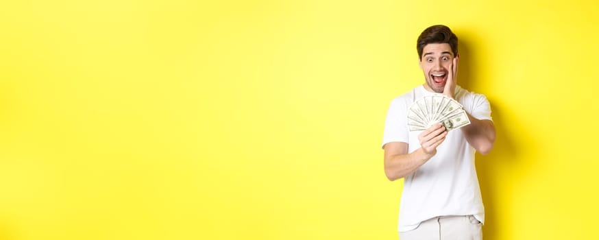 Man looking amazed at money, winning cash prize, standing against yellow background.