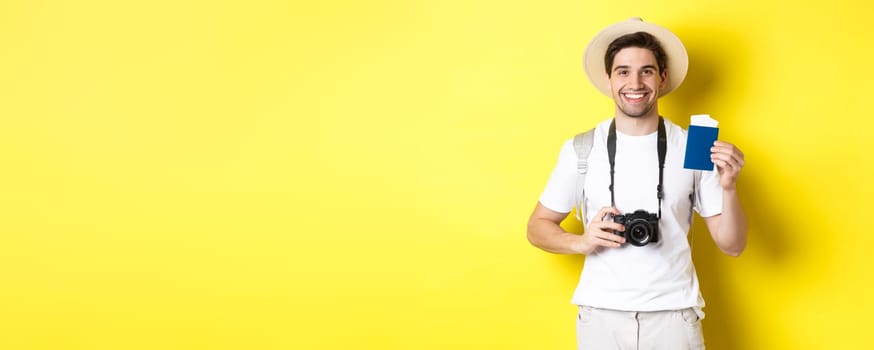Travelling, vacation and tourism concept. Smiling man tourist holding camera, showing passport with tickets, standing over yellow background.