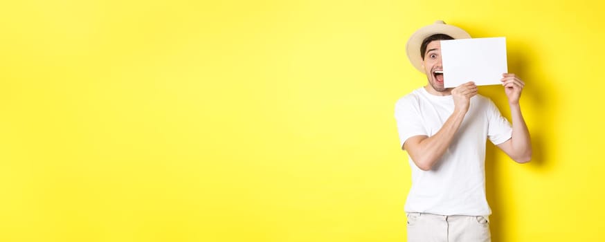 Excited tourist on vacation showing blank piece of paper for your logo, holding sign near face and smiling, standing against yellow background.