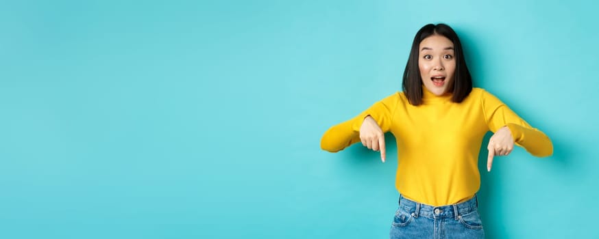 Shopping concept. Surprised attractive asian girl showing advertisement on blue background, pointing fingers down, stare at camera and say WOW.