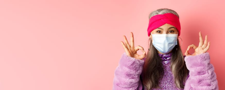 Covid-19, virus and fashion concept. Close-up of fashionable asian senior woman in face mask, showing OK signs and looking happy, pink background.