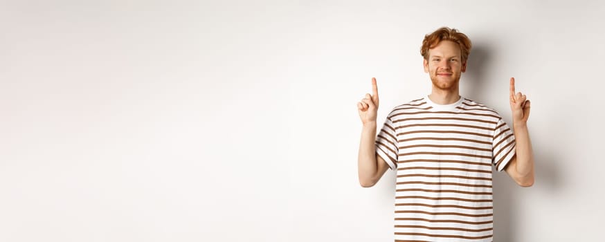 Smiling redhead guy with beard, pointing fingers up and showing advertisement, standing over white background.