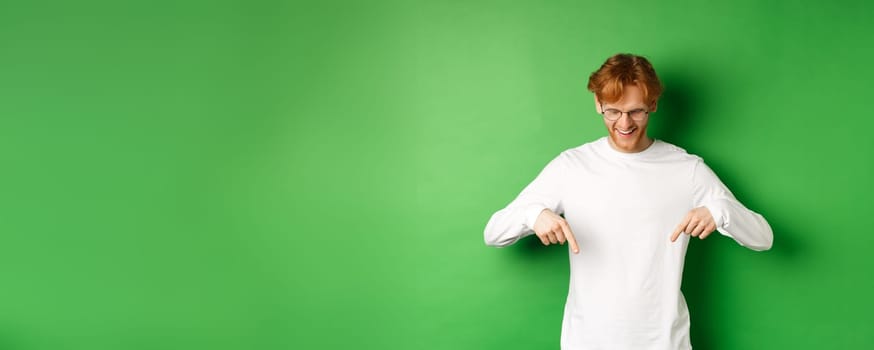 Handsome redhead man in glasses and white long-sleeve checking out advertisement, pointing fingers and looking down with pleased smile, standing over green background.