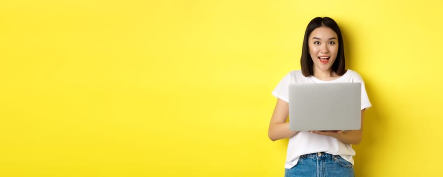 Cheerful asian woman studying on laptop and smiling, standing in white t-shirt and jeans against yellow background.