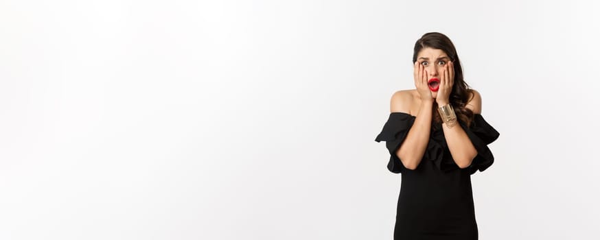 Fashion and beauty. Shocked and worried woman in black dress looking scared, standing anxious over white background.