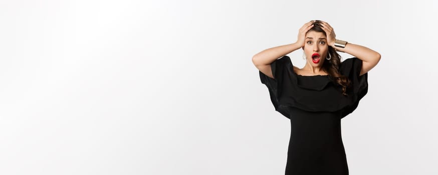 Fashion and beauty. Frustrated young woman in black dress, gasping and holding hands on head in panic, standing worried against white background.