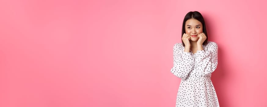 Cute and shy asian girl blushing, touching cheeks and looking left at copy space silly, standing against pink background.