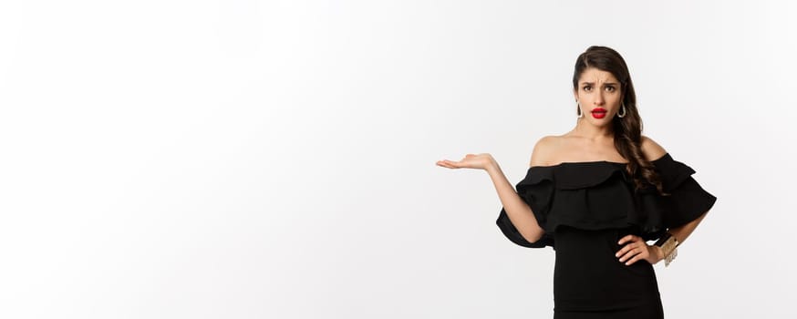 Fashion and beauty. Annoyed woman in black dress raising hand, so what gesture, looking confused at camera, standing bothered over white background.