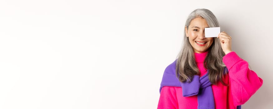 Shopping concept. Stylish asian senior woman smiling and showing plastic credit card, paying contactless, standing over white background.