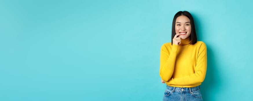 Image of good-looking asian woman in stylish outfit, touching lip and smiling at camera with happy face, standing over blue background.