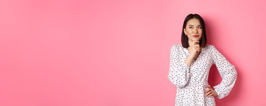 Thoughtful happy asian woman making decision, smiling satisfied and looking at upper left logo, thinking or choosing, standing in dress against pink background.