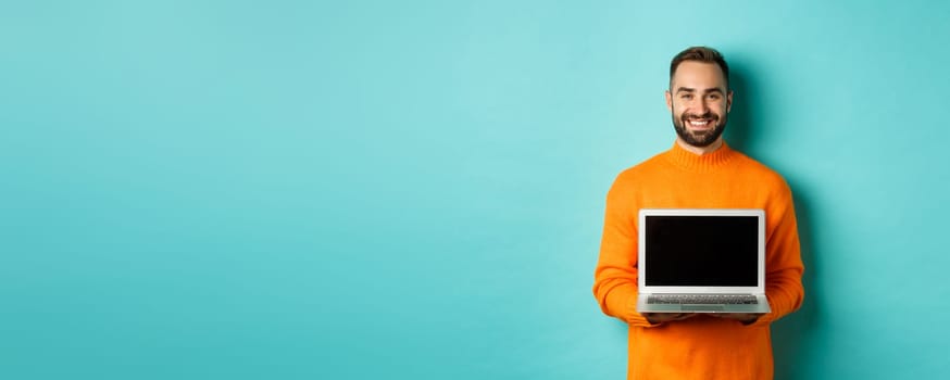 Handsome bearded man in orange sweater showing laptop screen, demonstrating online store, standing over light blue background.