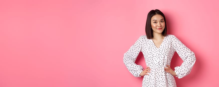 Beautiful korean lady in dress looking at camera and smiling, waiting for something, standing against pink background.