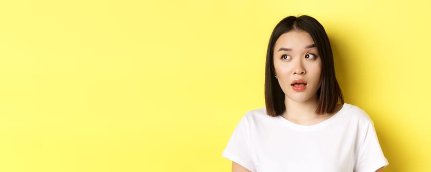 Close up of beautiful asian woman with clean perfect skin, raising eyebrow and looking left with intrigued face, standing over yellow background.