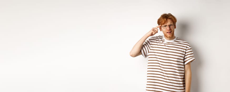 Annoyed young man with red hair scolding person for being stupid, pointing at head and staring at camera, white background.