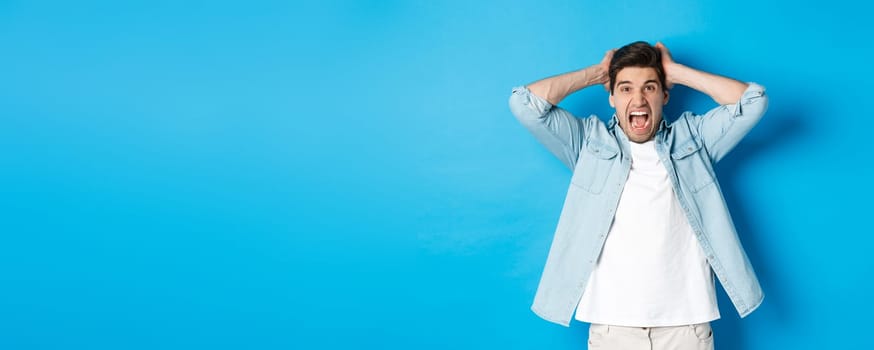 Frustrated guy losing, yelling displeased and looking at camera, holding hands on head, standing over blue background.